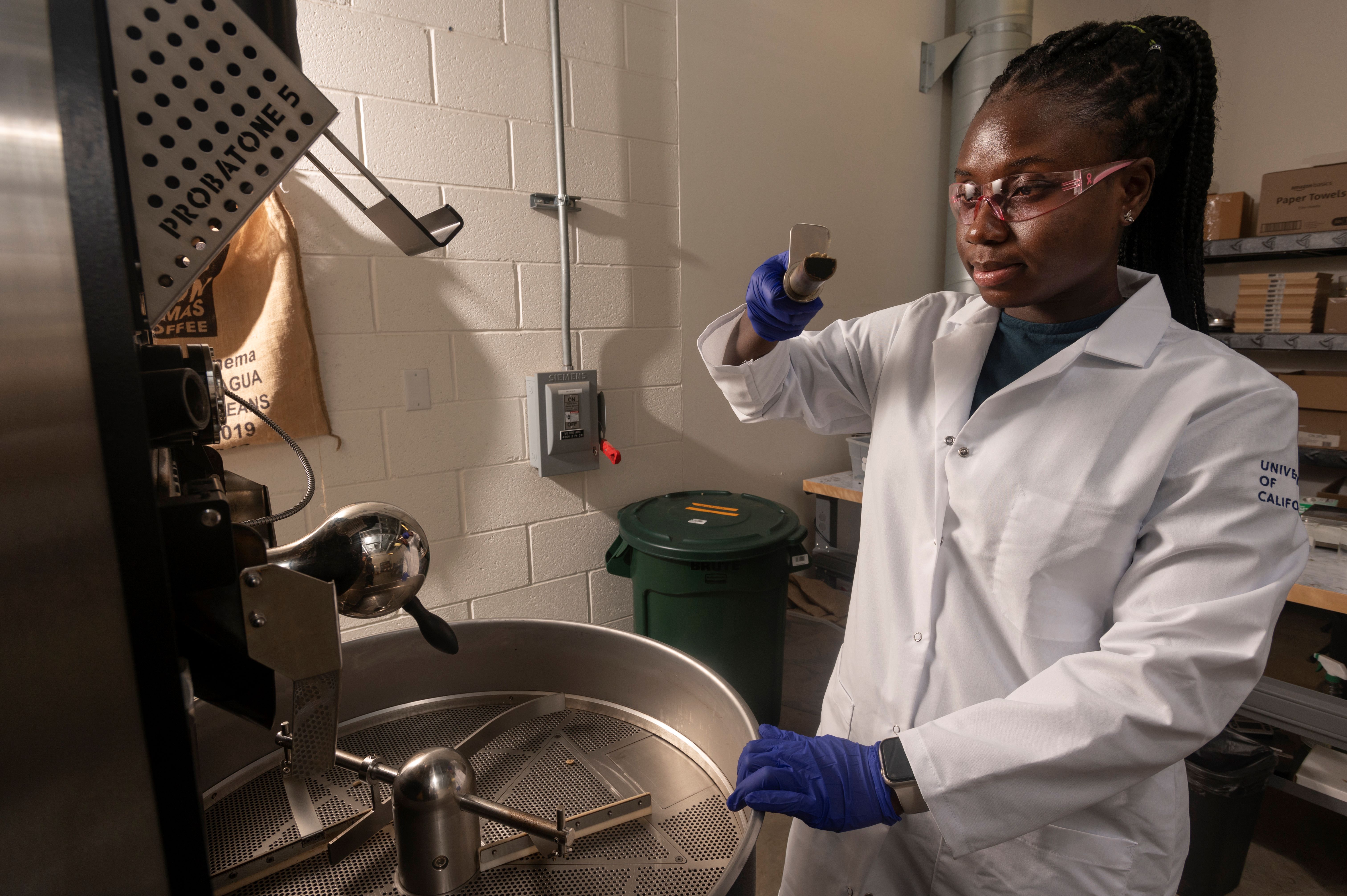 A black female researcher wearing a white lab coat and blue latex gloves removes a sample of coffee beans from a Probat coffee roaster