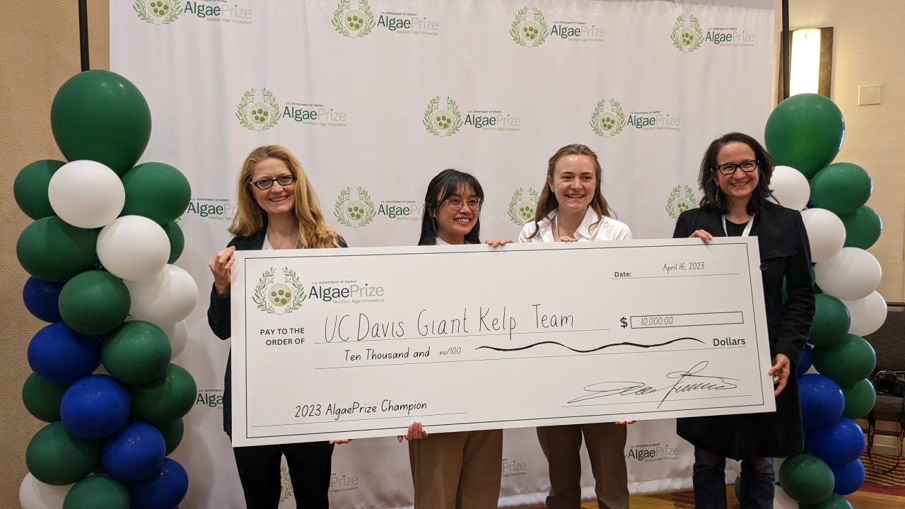 Two UC Davis students and two professors hold big check up in front of a step and repeat with AlgaePrize logo in green and white