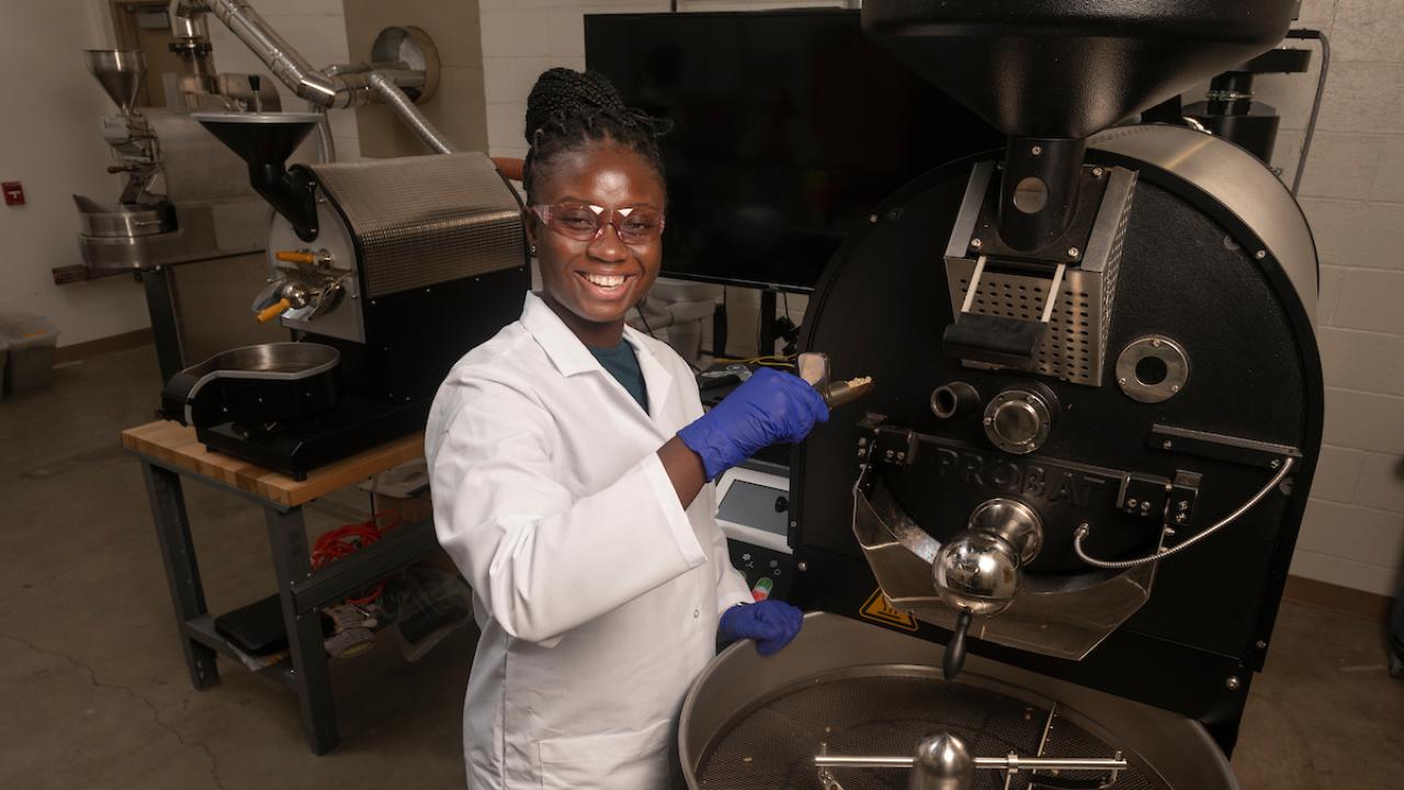 Laudia Anokye-Bempah stands in front of a coffee roaster. (Gregory Urquiaga/UC Davis)Laudia Anokye-Bempah stands in front of a coffee roaster