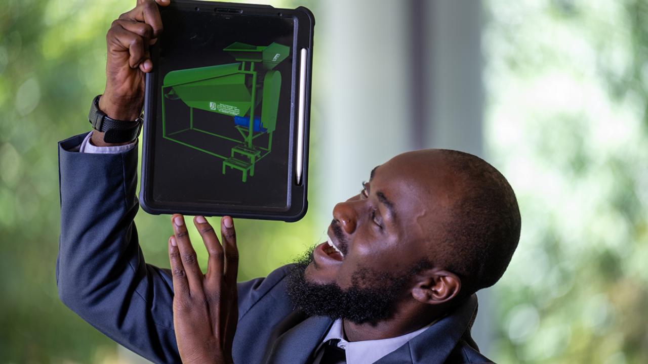 alt="Ismael Mayanja wearing a suit holds up a tablet with a green model displayed"