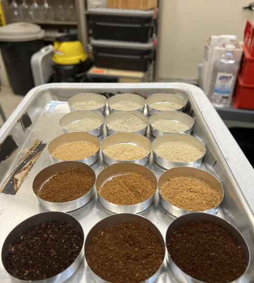 Three rows of ground coffee samples of different colors are lined up on a metal tray