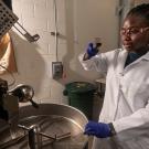 A black female researcher wearing a white lab coat and blue latex gloves removes a sample of coffee beans from a Probat coffee roaster